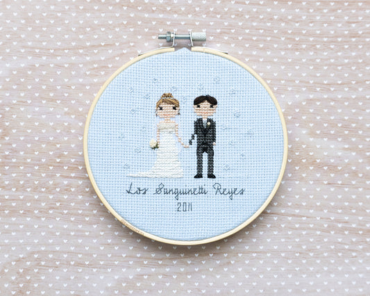 A cross-stitched bride and groom with subtle bubbles hand-stitched around them. It's framed in a wooden embroidery hoop and reads "Los Sanguinetti Reyes"