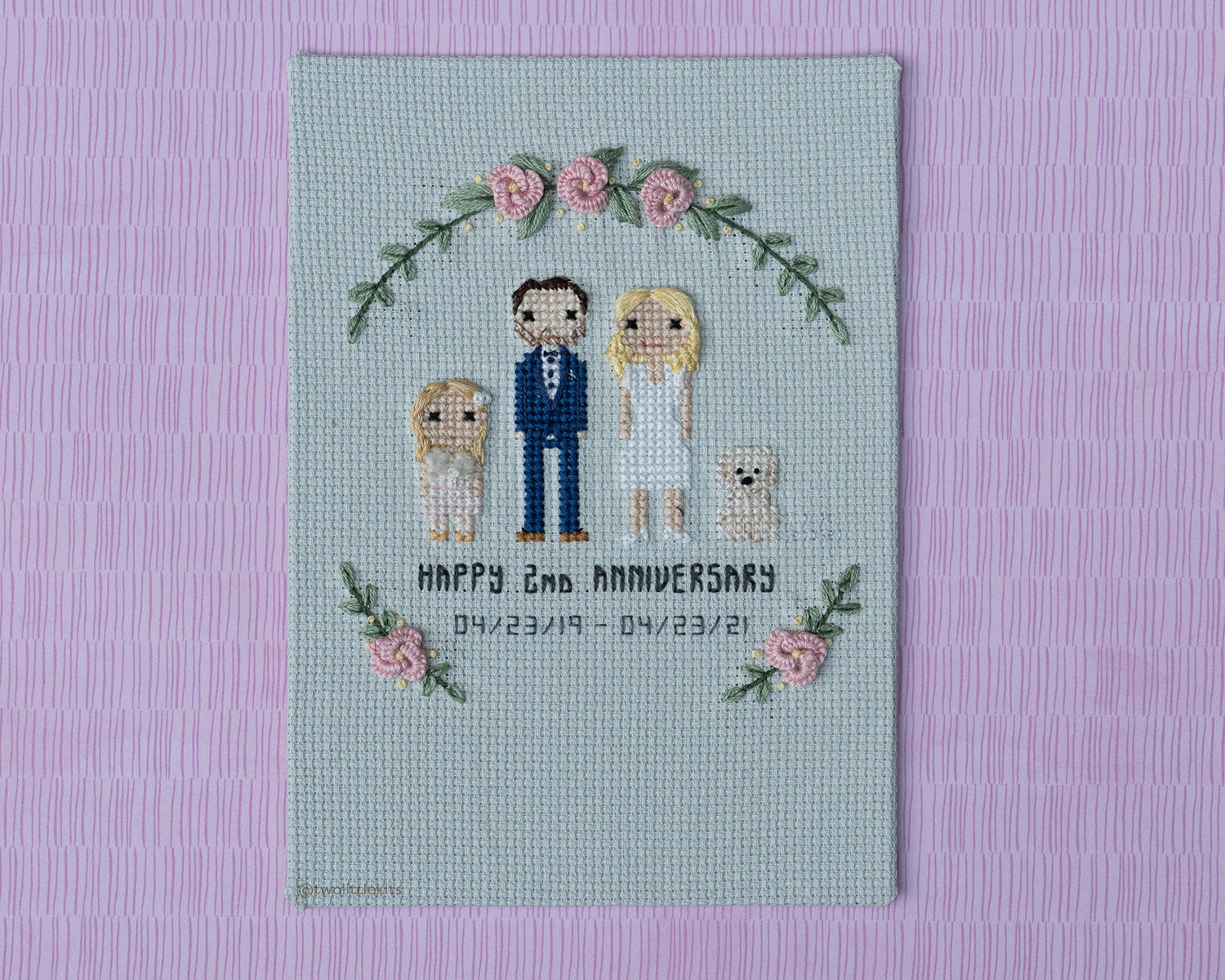 A cross-stitched bride and groom with their daughter and dog. It is mounted on a rectangular board and reads "HAPPY 2nd ANNIVERSARY" with two dates. Around everything is hand-stitched florals.
