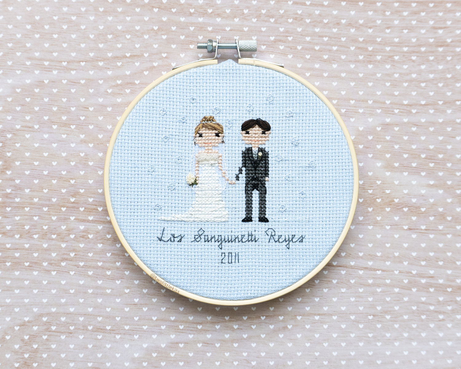 A cross-stitched bride and groom with subtle bubbles hand-stitched around them. It's framed in a wooden embroidery hoop and reads "Los Sanguinetti Reyes"