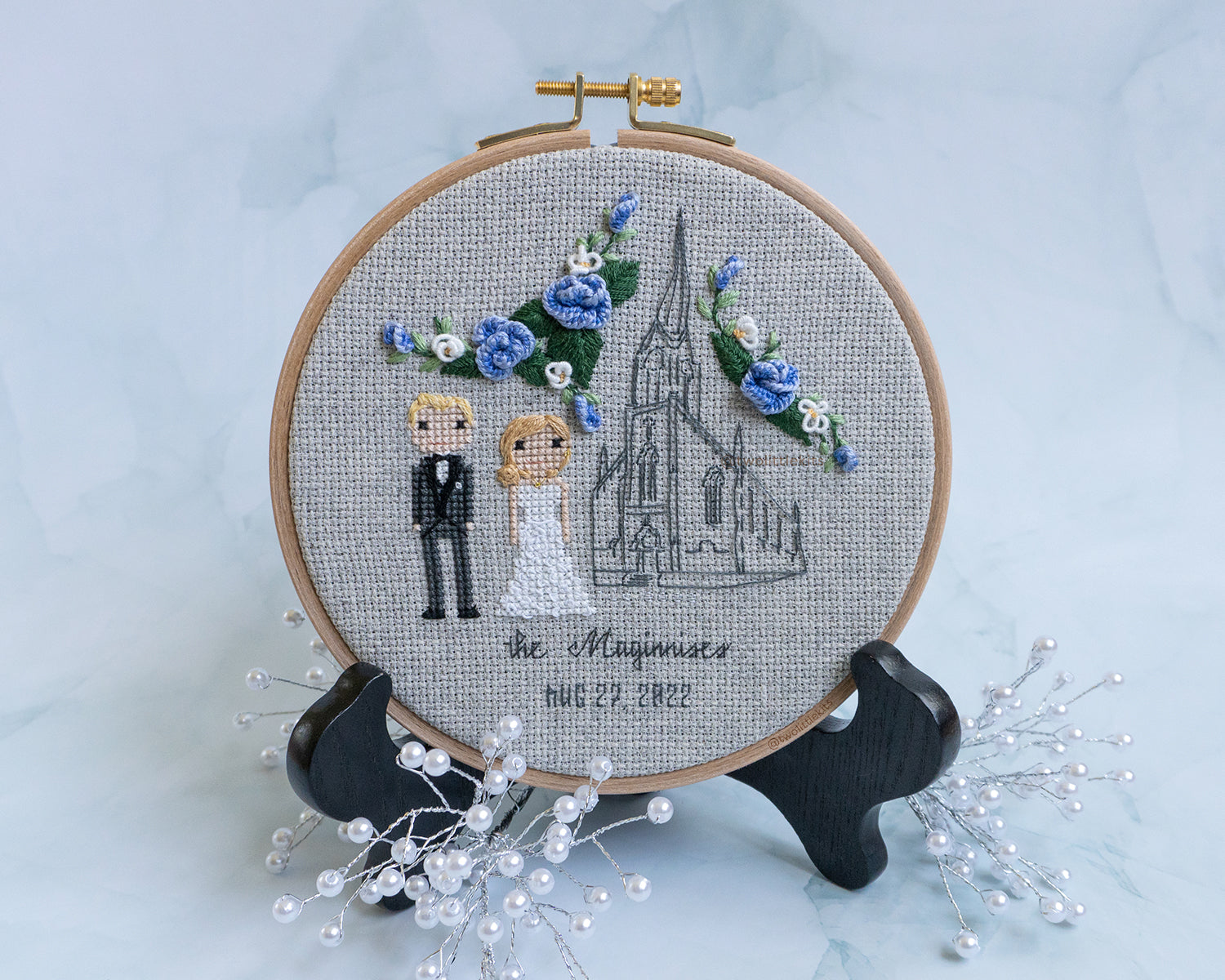 A cross-stitched bride and groom next to an outlined church building. In the "white space" there is blue and white hand stitched florals. The piece is framed in a wooden embroidery hoop and reads "The Maginises"