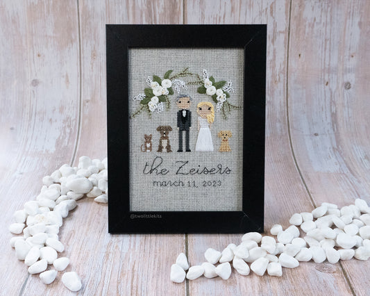A cross-stitched bride and groom with floral arrangement around them and their three pets; two dogs and one cat. It is framed in a black wooden frame and reads "The Zeisers"